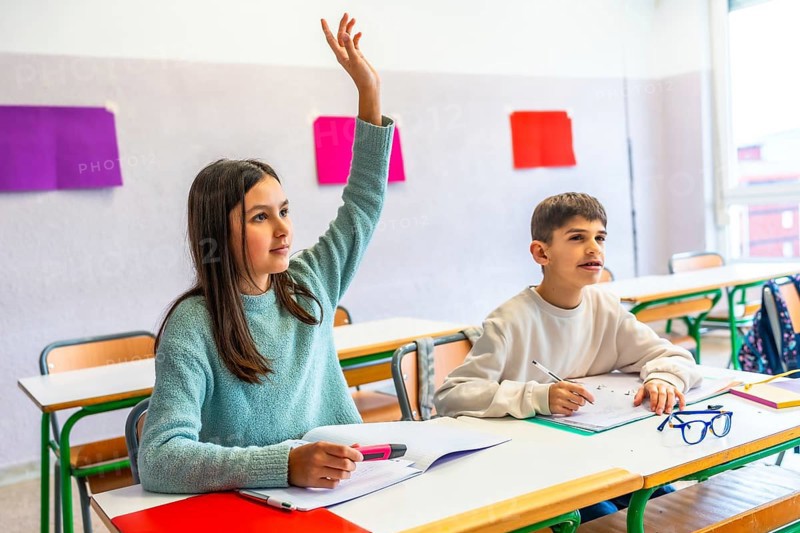 Girl raising hand to answer a question during class in elementary school while her classmate is taking notes