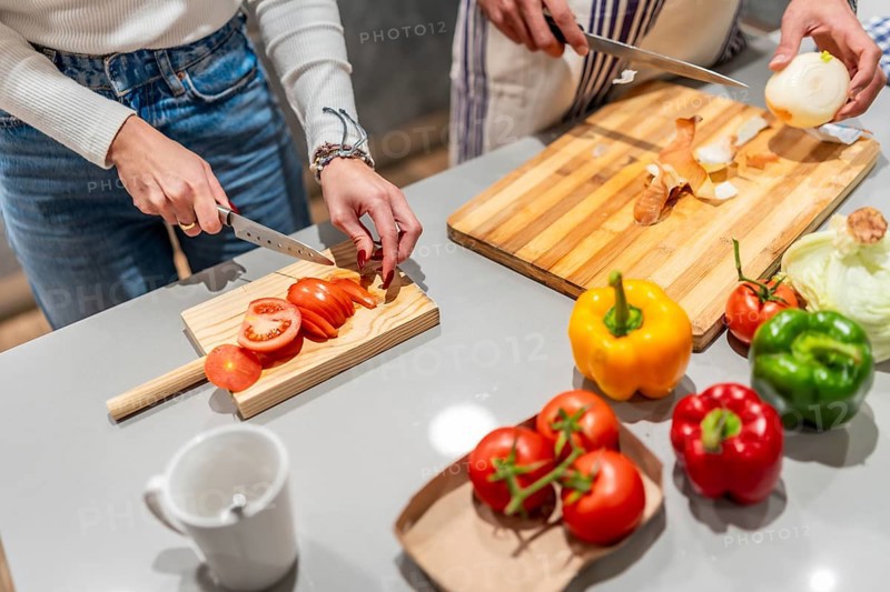 Two chefs slicing fresh tomatoes and onions on wooden cutting boards, surrounded by colorful bell peppers in a modern kitchen