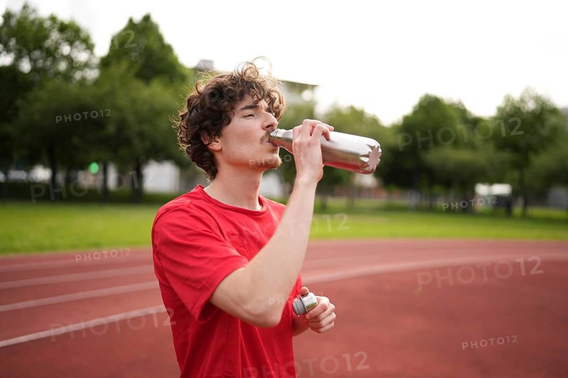 Thirsty caucasian young amle athlete drinking water in a reusable bottle after training in a running track
