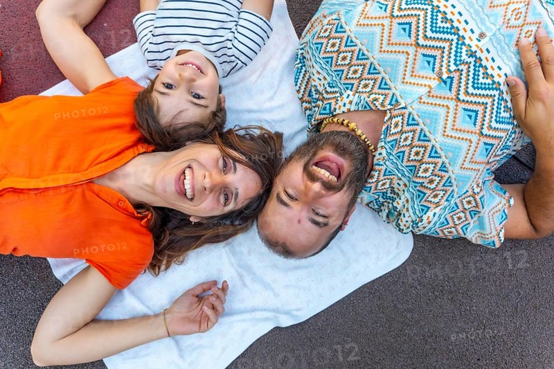 A family of three, a man, a mother and a son, are laying on a blanket on the ground. The man is wearing a blue and white shirt with a pattern of diamonds. The children are smiling