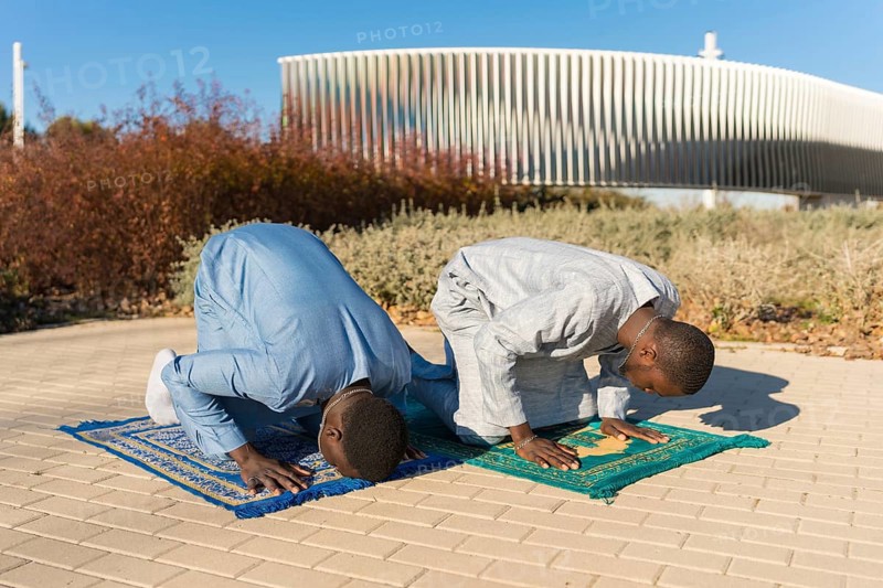 Two senegalese men wearing traditional dashiki clothing are kneeling on prayer rugs, performing salat during the holy month of ramadan