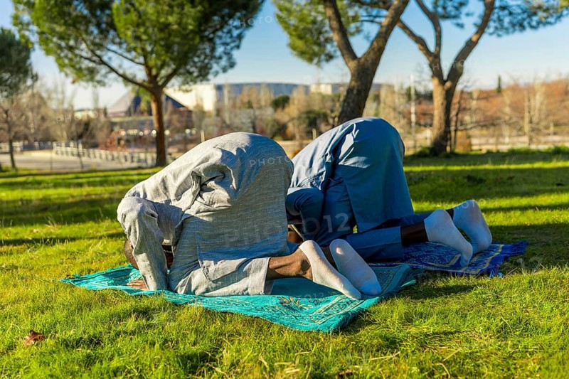 Two african muslim men are kneeling in prayer on prayer rugs in a park, wearing traditional clothing