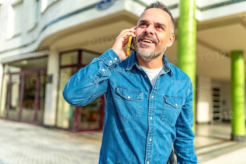 Happy mature man with denim clothes smiling talking to the mobile in the street