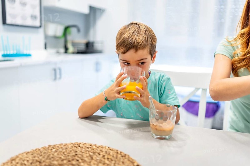 School boy drinking orange juice at home with his mother at the side before going to school