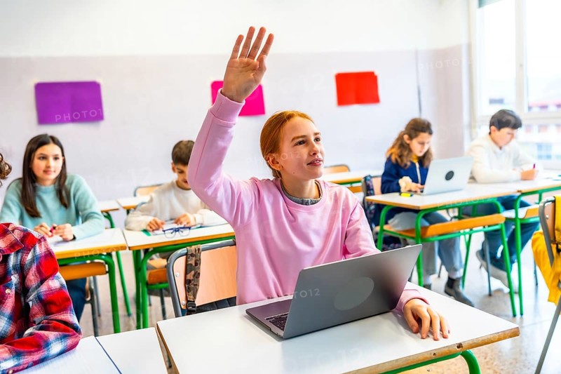 Smart student raising hand to answer teacher's question in modern classroom with laptops and focused classmates