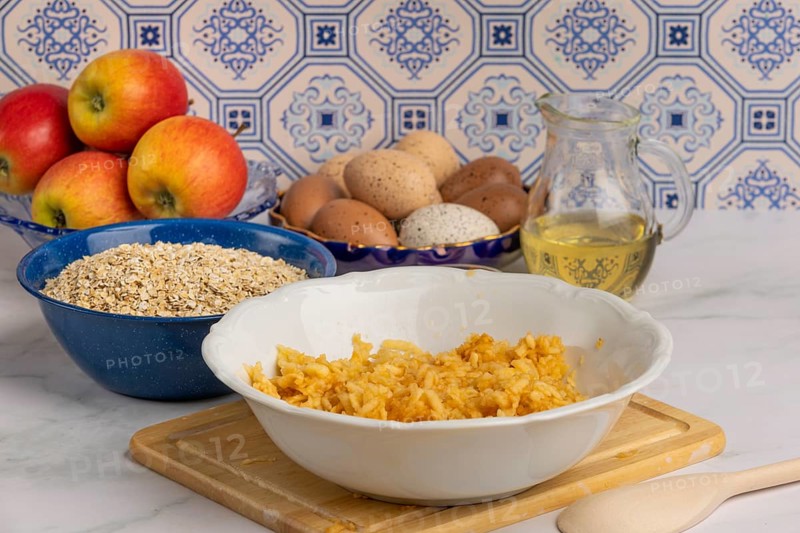 Grated apple in a bowl in front of oat flakes, eggs and honey
