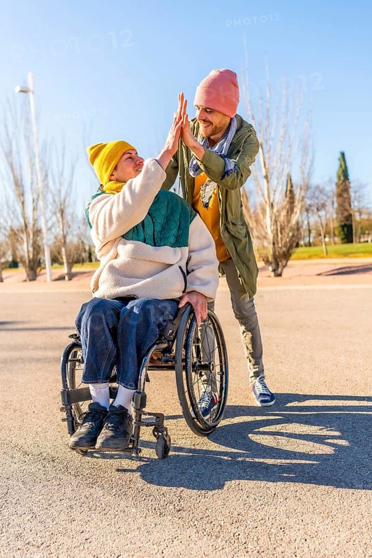 Two friends celebrating something with a high five in a park, one of them using a wheelchair, concept of disability and friendship