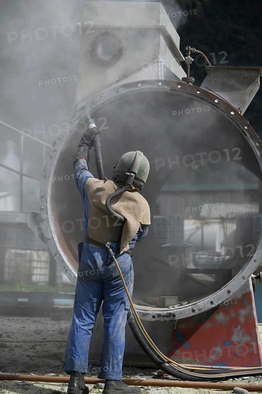 A man weating full safety gear uses ground glass to sandblast the steel casing of a meat drying machine for a rendering plant