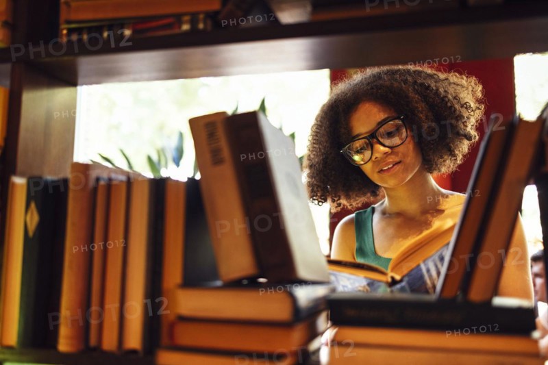 Happy African American female student studing in university library and looking for the book, wearing glasses