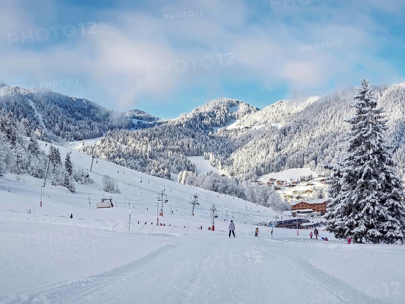 Picturesque snowy mountain landscape with slopes, lifts, and skiers at La Clusaz ski resort, Annecy, France. Snow covered woods and a small village with cabins nestled in the valley