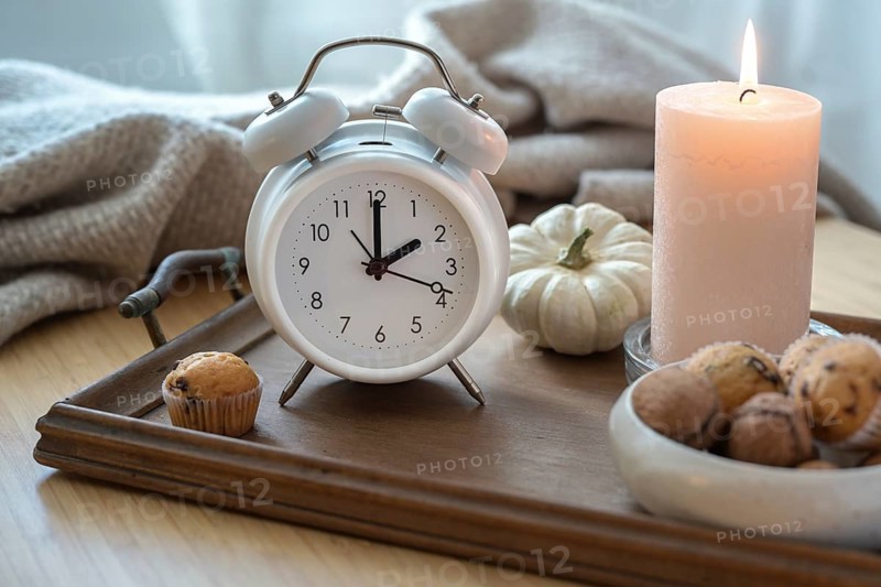 White vintage alarm clock, candle, autumn decoration and sweets on a nightstand with a natural woolen blanket, fall back concept after daylight saving time, selected focus
