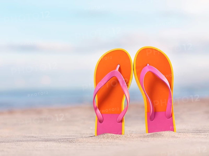 Close up of a pair of colorful orange flip flops in sand at the beach under blue sky background