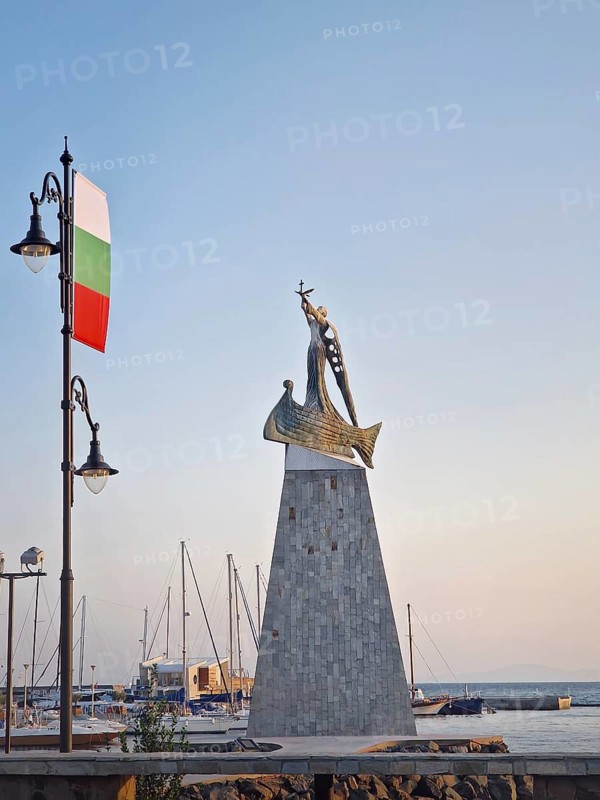 The statue of Saint Nicholas patron of sailors in the old town of Nessebar, Bulgaria. Serene sunset at harbor with boats docked in calm waters, a street lamp with adorned bulgarian flag in foreground