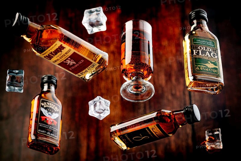 Four whisky bottles and a glass of whisky with floating ice cube cubes against a wooden background