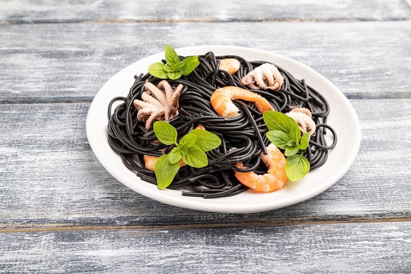Black cuttlefish ink pasta with shrimps or prawns and small octopuses on gray wooden background. Side view, close up