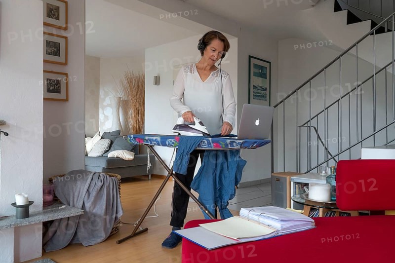 Woman, mid-50s, works from home, with laptop and communicates with colleagues via headset, home office, irons laundry in parallel