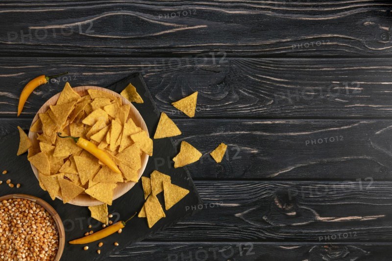 Bowl of tortilla chips next to raw sweetcorn and yellow peppers on a dark wooden table