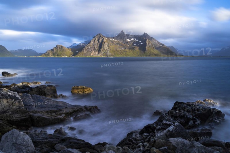 Landscape with sea and mountains on the Lofoten Islands, view across the fjord to the small town of Flakstad and the mountain Flakstadtinden as well as other mountains. Rocks in the foreground. At night at the time of the midnight sun in good weather, some clouds in the sky. Long exposure, blurred clouds. Early summer. Flakstadoya, Lofoten, Norway, Europe