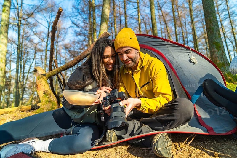 Couple of hikers reviewing photos on professional camera while having a meal in front of their tent during camping trip in forest