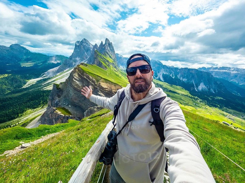 Photographer taking a selfie with outstretched arm, capturing the breathtaking seceda peaks and lush valleys of the dolomites during a sunny summer day