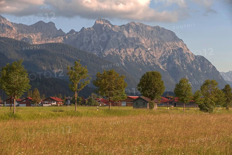 View of a village in front of majestic mountains, surrounded by green meadows and trees under a cloudy sky, Wallgau, Bavaria, Germany, Europe