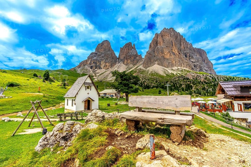 Picturesque landscape featuring a wooden bench, a small chapel, and the majestic peaks of the sella group in the italian dolomites during summer