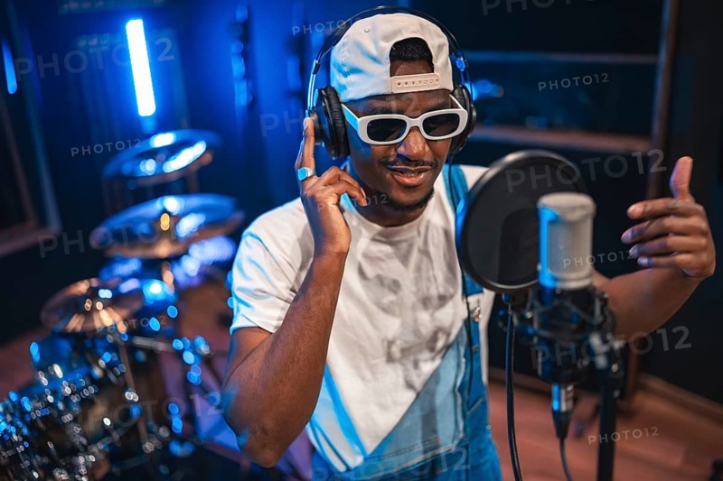 Young fashionable artist wearing headphones and sunglasses, singing into a microphone and recording a song or album in a music studio