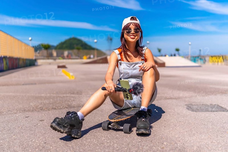 Young female asian influencer sitting on skateboard park holding mobile phone under the sun