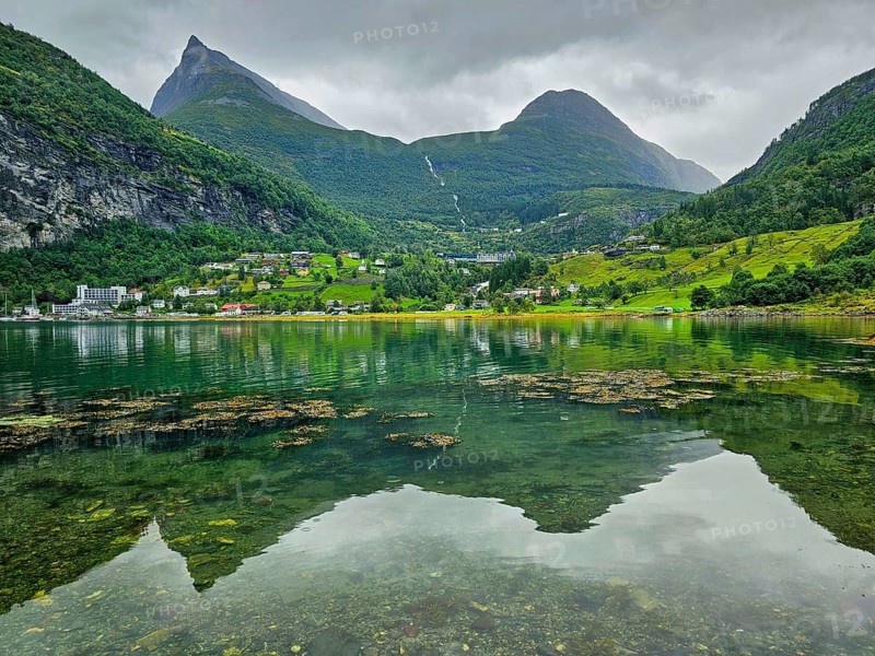 Mountainous landscape with a calm fjord reflecting the surrounding mountains and village, surrounded by green nature and under a cloudy sky, Geiranger, Geiranger Fjord, Stranda, Romsdal, Norway, Europe