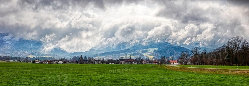 Green meadow and small village in the foreground, dramatic clouds, mountains in the background, Bad Feilnbach