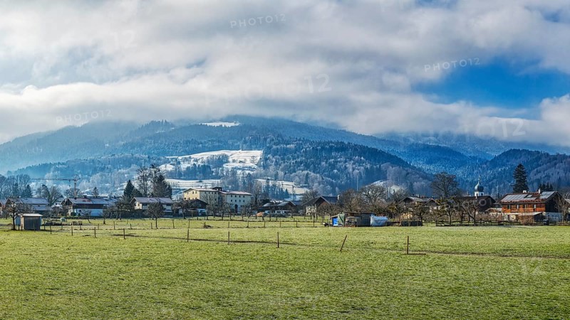 A village in front of snow-covered mountains under a cloudy sky on a green meadow, Bad Feilnbach, winter