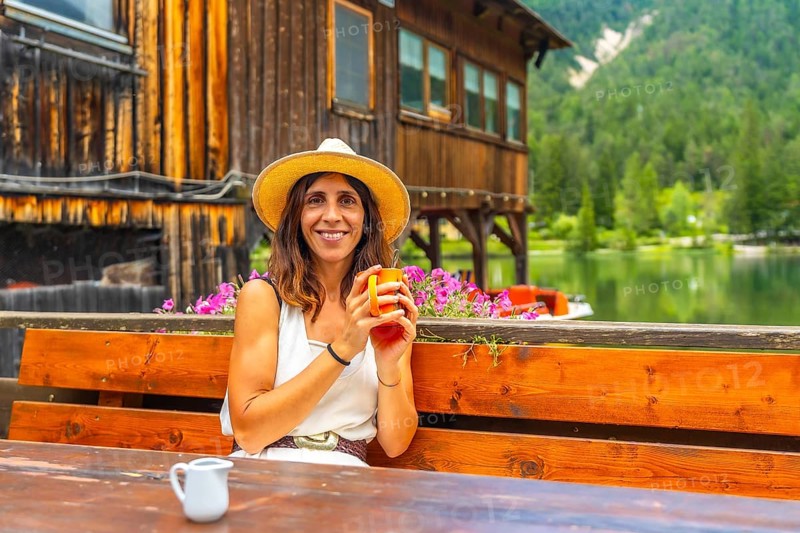Woman with straw hat enjoying a hot drink in a bar with mountain lake view in the dolomites