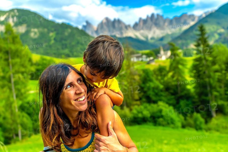 Mother carrying her son on her shoulders, enjoying a sunny day in the italian dolomites with the church of st. Magdalena nearby