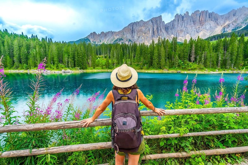 Female tourist with backpack admiring the stunning turquoise waters of lake carezza and the latemar mountain range in the italian dolomites during summer