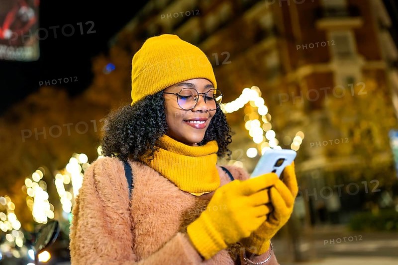 Smiling young woman wearing yellow winter clothes using smartphone in a city street at night with christmas lights