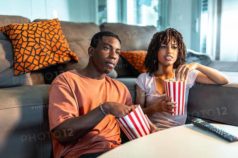 Young black couple enjoying a movie night at home, sitting on a comfortable sofa, eating popcorn from striped buckets, and watching television