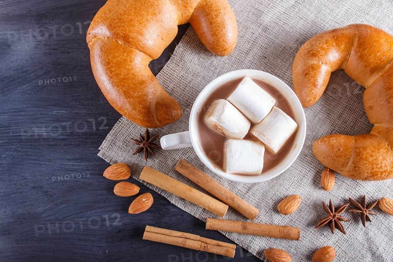 A cup of hot chocolate with marshmallow, croissants and spices on linen napkin and black wooden background. top view, copy space