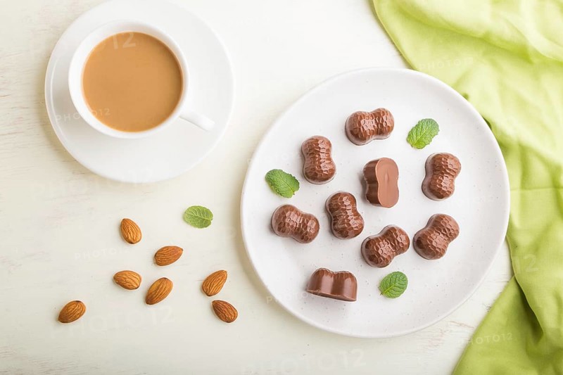 Chocolate candies with almonds and a cup of coffee on a white wooden background and green textile. top view, flat lay, close up