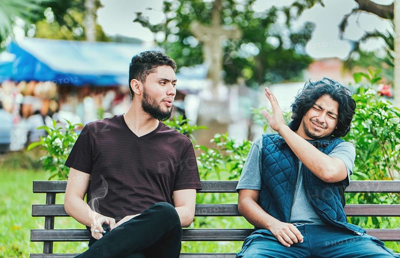 Man smoking and exhaling smoke to another person near him. Man smoking cigarette near people in public