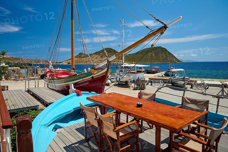 A harbour scene with wooden boats and a table with chairs and a blue boat in the foreground in sunny weather, Patmos Marine, boatyard, Diakofti, Patmos, Dodecanese, Greek Islands, Greece, Europe
