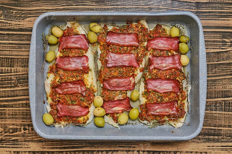 Roasted fish fillets are layered with bacon and herb sauce, accompanied by green olives. This delicious dish is prepared in a baking tray, perfect for sharing