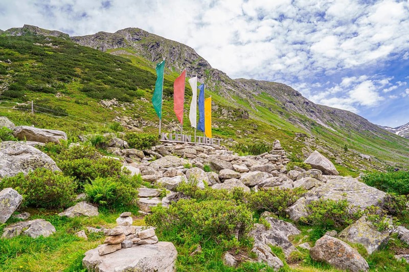 Alpine landscape with colourful flags and a sign next to piles of stones, green vegetation and cloudy sky, Klein Tibet, Zillertal, Austria, Europe