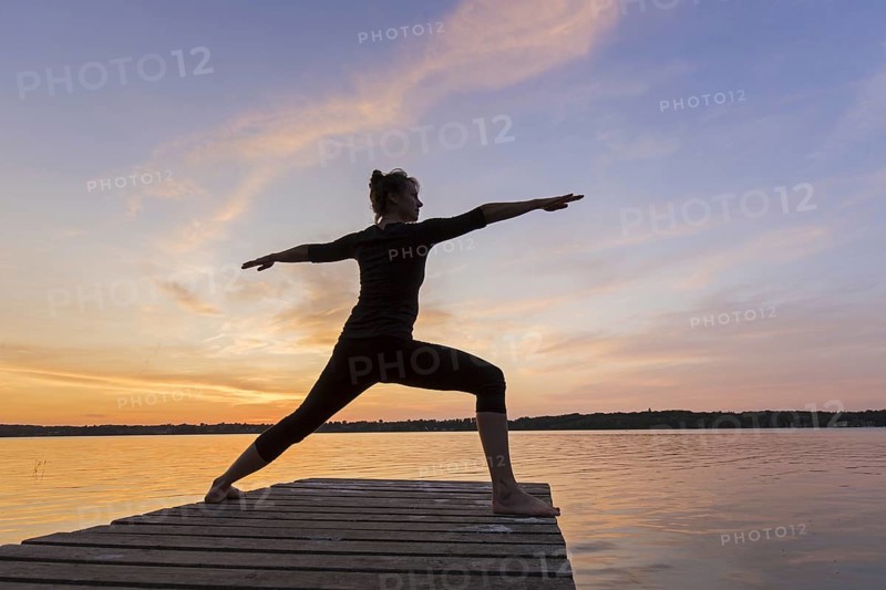 Woman practicing yoga posture Virabhadrasana II, Warrior Pose, lunging standing asana on jetty at lake at sunset