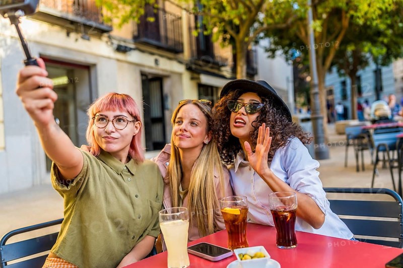 Three modern diverse women taking selfie with digital camera and tripod at the terrace of a bar