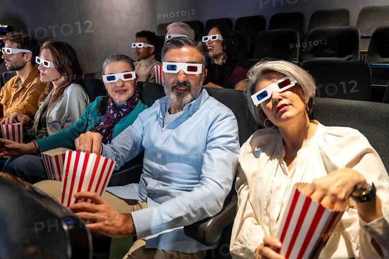 People of different ages and ethnicities wearing 3d glasses and eating popcorn while watching a movie in a cinema