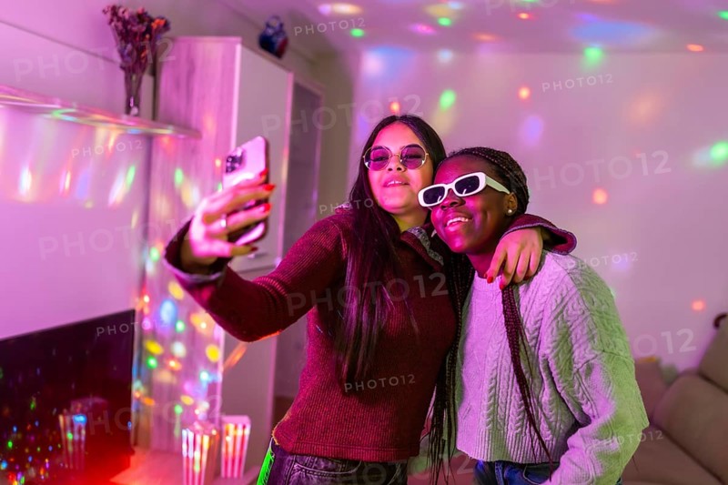 Two teenage girls wearing sunglasses are taking a selfie with a smartphone at a home party with disco lights