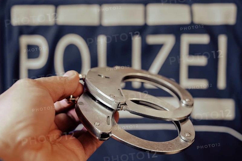 A pair of handcuffs is held by one hand, a police sign in the background