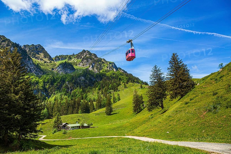 The Hochfelln cable car travels over green meadows and fir trees against a backdrop of impressive mountains and blue skies in Chiemgau, Bergen, Bavaria, Germany, Europe