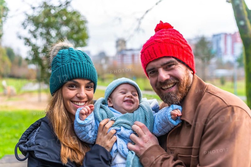 Smiling parents holding their laughing baby wearing winter clothes, enjoying a sunny winter day at the park