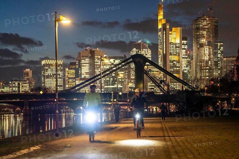 Skyline of the city centre of Frankfurt am Main, cyclist with light on the cycle path, pavement, promenade along the river Main, dusk, Hesse, Germany raft bridge in the background